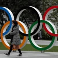 A woman wearing a protective face mask walks past the Olympic rings in front of the Japan Olympics Museum in Tokyo on March 30. | REUTERS