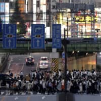 Pedestrians cross a road at dusk on Friday in Tokyo\'s Shinjuku Ward, where a cluster of coronavirus infections has been detected. | BLOOMBERG