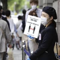 An employee holds a board reminding customers to maintain social distancing at the entrance of a department store in Kobe on May 22. | KYODO

