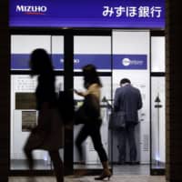 Pedestrians walk past a customer using an automated teller machine at a branch of Mizuho Bank in Tokyo in May. | BLOOMBERG