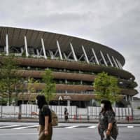 People walk past the National Stadium, the main venue for the 2020 Olympic and Paralympic Games now postponed due to the coronavirus pandemic, in Tokyo on Sunday. | AFP-JIJI