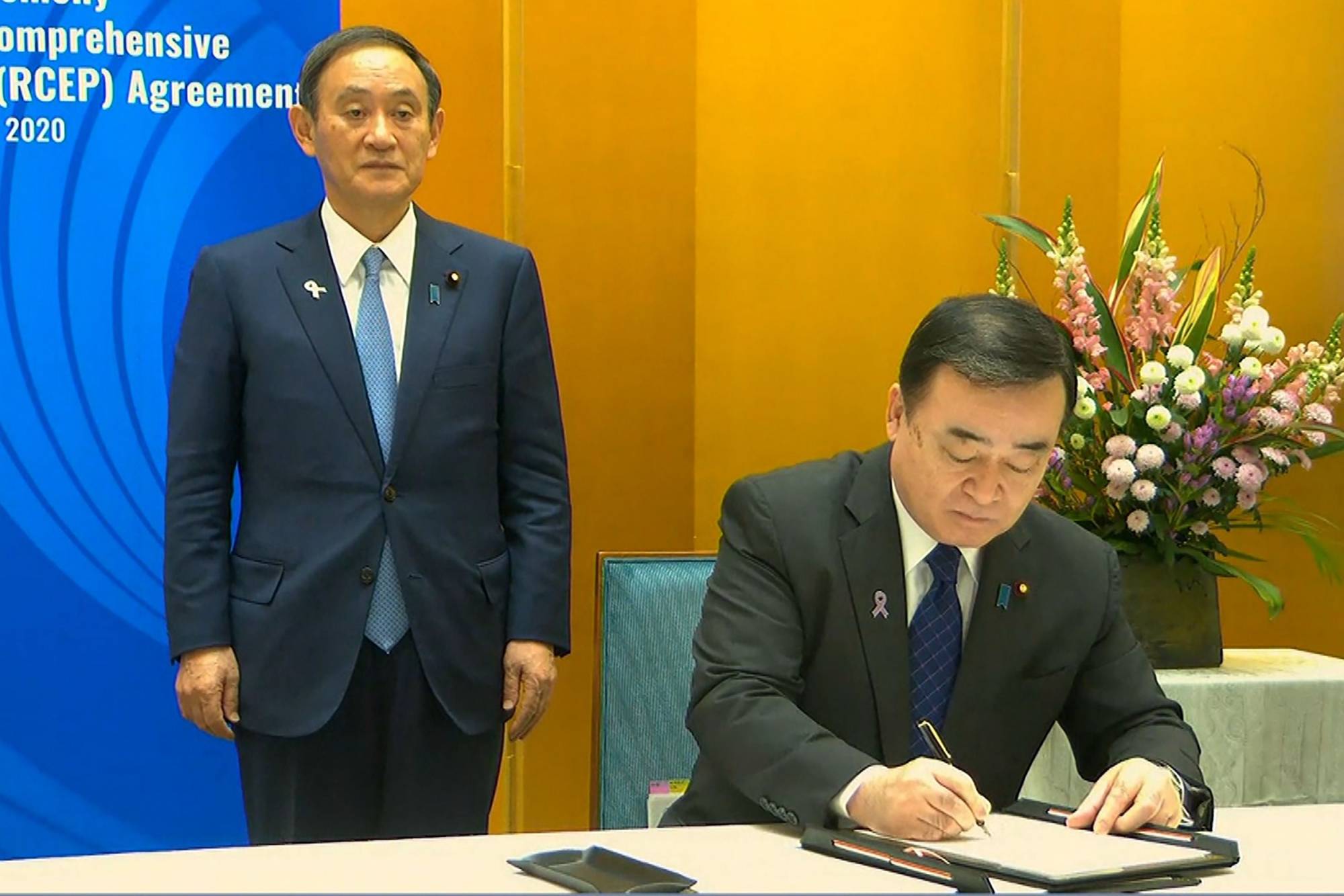 A screen grab taken from Vietnam Host Broadcaster's on Sunday shows Japan's Prime Minister Yoshihide Suga watching Minister of Economy, Trade and Industry Hiroshi Kajiyama sign the Regional Comprehensive Economic Partnership trade pact. | VIETNAM HOST BROADCASTER / VIA AFP-JIJI 