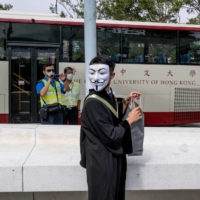 A graduating student wearing a Guy Fawkes mask attends a protest at the Chinese University of Hong Kong as a campus security officer observes with a video recorder on Thursday. | BLOOMBERG
