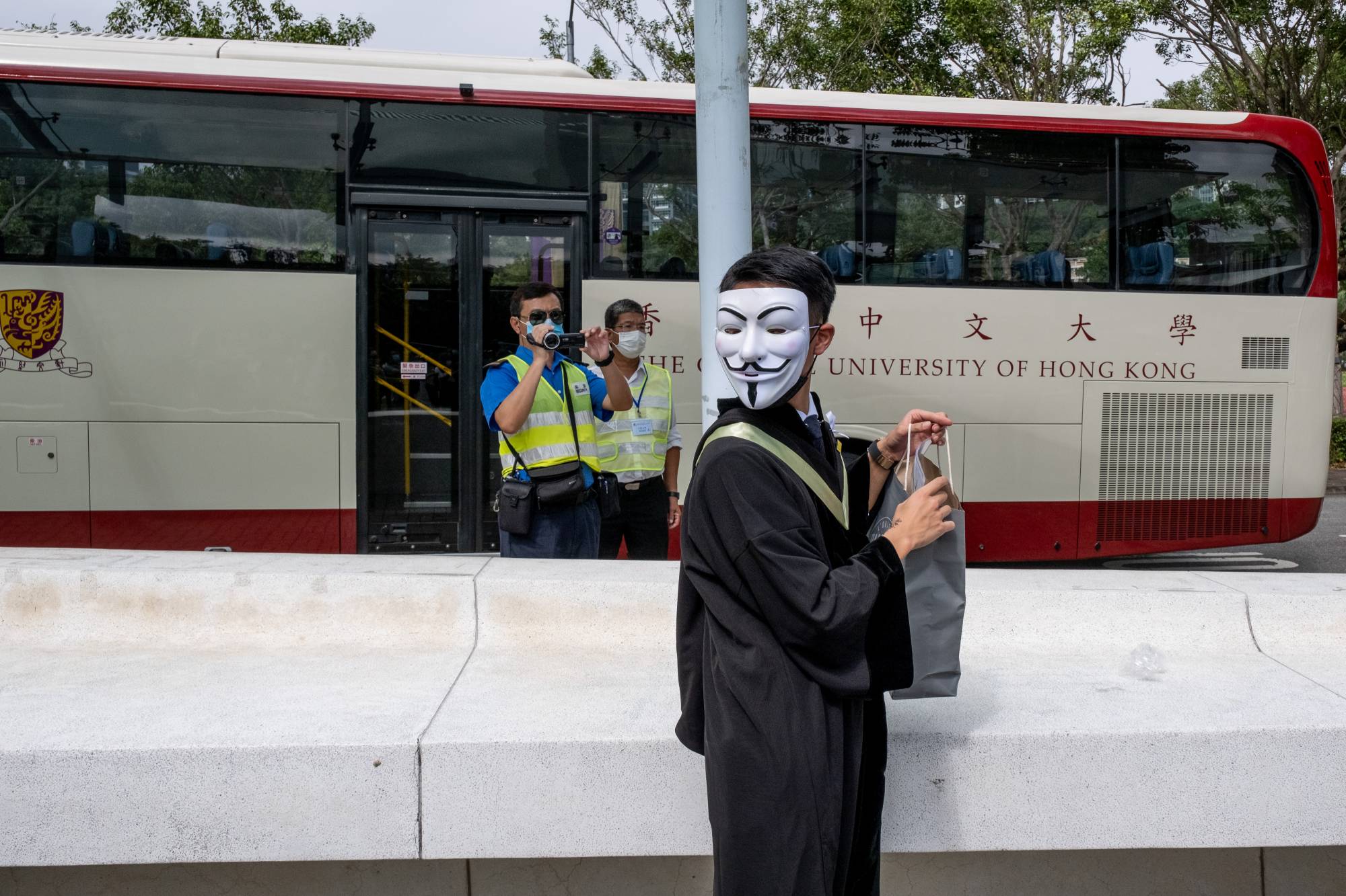 A graduating student wearing a Guy Fawkes mask attends a protest at the Chinese University of Hong Kong as a campus security officer observes with a video recorder on Thursday. | BLOOMBERG