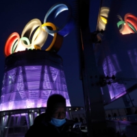 A man wearing a face mask walks near the lit-up Olympic rings at the top of the Olympic Tower, a year ahead of the opening of the 2022 Winter Games, in Beijing on Feb. 4. | REUTERS