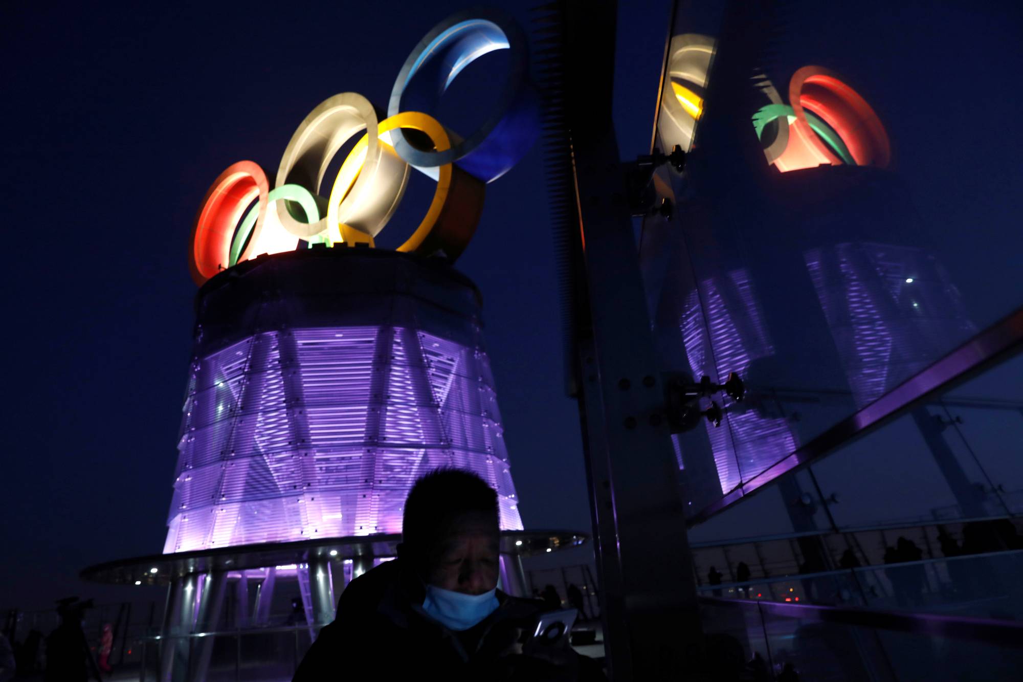 A man wearing a face mask walks near the lit-up Olympic rings at the top of the Olympic Tower, a year ahead of the opening of the 2022 Winter Games, in Beijing on Feb. 4. | REUTERS