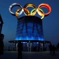 People wearing face masks walk near the lit-up Olympic rings at the top of the Olympic Tower, a year ahead of the opening of the 2022 Winter Games, in Beijing on Feb. 4. | REUTERS