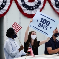 People hold flags and a sign as they attend a Democratic National Committee drive-in rally to celebrate U.S. President Joe Biden\'s 100th day in office, in Duluth, Georgia, on Thursday. | REUTERS