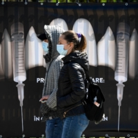 People wearing masks walk by a banner showing syringes as vampire fangs at a vaccination center at Bran Castle  — purported to be an inspiration for the vampire\'s towering home in Bram Stoker’s novel \"Dracula\"  — in Bran, Romania, on Saturday. | AFP-JIJI