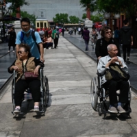 An older man and woman are pushed in wheelchairs along a street in Beijing on Tuesday.  | AFP-JIJI
