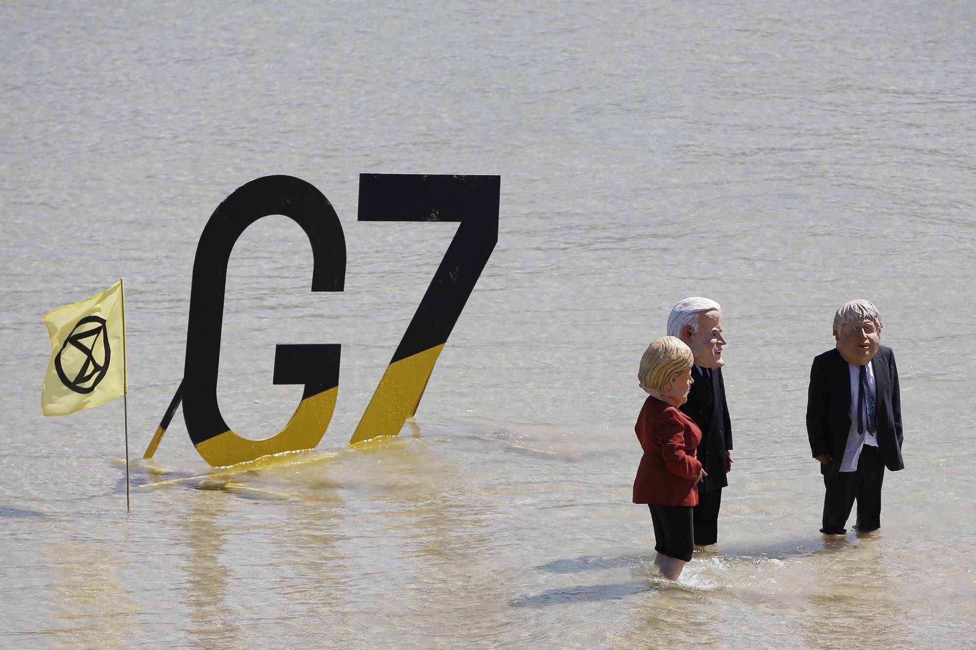 Extinction Rebellion protesters stand in front of a Group of Seven sign wearing masks depicting German Chancellor Angela Merkel, U.S. President Joe Biden and British Prime Minister Boris Johnson during the G7 leaders summit in St. Ives, England, on Sunday. | BLOOMBERG