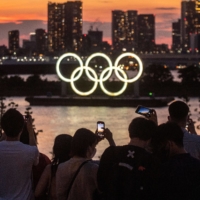 People take pictures as the Olympic rings are lit up at dusk on Tokyo\'s Odaiba waterfront area. The Tokyo Olympic organizing committee on Saturday reported 21 more COVID-19 cases related to the Games. | AFP-JIJI