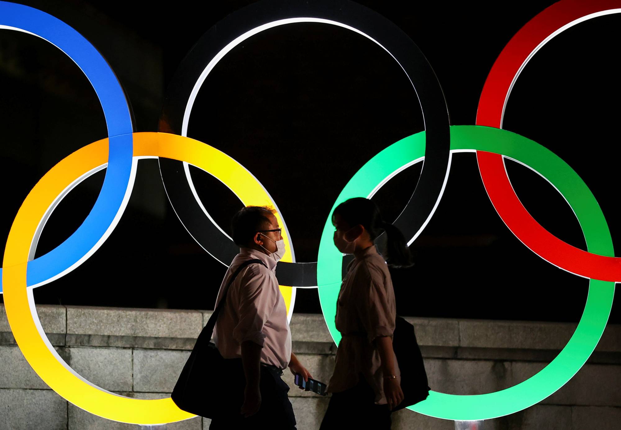 People wearing protective face masks walk past an illuminated Olympic rings monument during the Tokyo Games in the capital on Thursday. | REUTERS