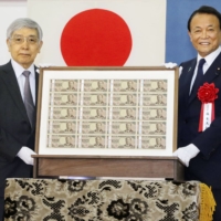Bank of Japan Gov. Haruhiko Kuroda (left) and Finance Minister Taro Aso show redesigned ¥10,000 bank notes printed on Thursday in Tokyo. | NATIONAL PRINTING BUREAU / VIA KYODO