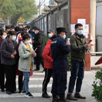 People wait in line to receive COVID-19 vaccine booster shots as a security guard checks the body temperature of a man along a street in Beijing on Saturday. | AFP-JIJI