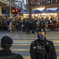 Police stand guard as pedestrians wait to cross a road near a flower market in Hong Kong on Sunday. Hong Kong denied that criticism of the city\'s strict policy to push for zero COVID-19 cases is illegal under a sweeping national security law that has silenced dissent in the former British colony. | BLOOMBERG