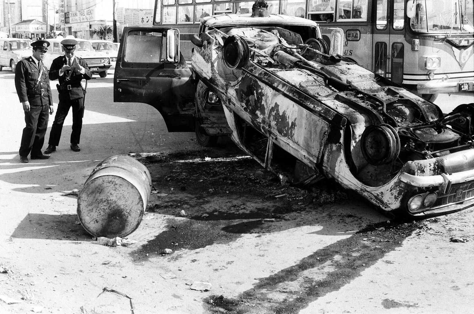 Police officers examine an upturned car that was damaged during riots in the streets of Koza, Okinawa, in December 1970. | HIROAKI YAMASHIRO