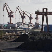 Piles of coal sit near port facilities as gantry cranes stand in the background at Qinhuangdao Port in 2016. China\'s efforts to quell surging coal prices showed signs they’re working, with benchmark prices dropping for the first time in a year as the country\'s production rose to the highest in seven months. | BLOOMBERG
