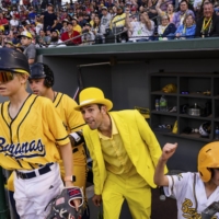 Jesse Cole, center in yellow tux, the owner of the Savannah Bananas, in the dugout for a road game with the Kansas City Monarchs, in Kansas City, May 7, 2022. TikTok choreography, dancing umpires, a ballet-trained first-base coach: This collegiate summer league team has amassed a big following by leaning into entertainment.  | SHAWN BRACKBILL / THE NEW YORK TIMES

