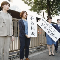 Plaintiffs\' lawyers hold signs, including one showing \"unjust ruling,\" in front of the Tokyo District Court on Thursday after a ruling on a damages claim by a sex business firm over a coronavirus cash handout snub. | KYODO