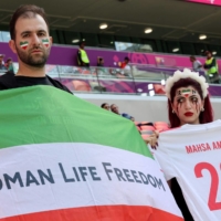 An Iran supporter with blood tears make-up on her face holds a soccer jersey with the name of Mahsa Amini, the Iranian woman who died at a hospital after been arrested by the country\'s morality police for violating Iran\'s strict dress code, poses with another supporter holding a protest flag as they attend the a 2022 World Cup Group B match between Wales and Iran in Al Rayyan, west of Doha, on Friday. | AFP-JIJI