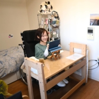 Yui Yuda sits in her Kyoto apartment, with a caregiver on standby near the entrance of the apartment. | NISHINIPPON SHIMBUN
