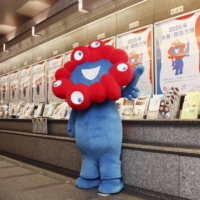 Myaku-Myaku, the mascot for the 2025 World Exposition in Osaka, poses for photos in April in front of posters for the Osaka Expo posted in Tokyo. | KYODO
