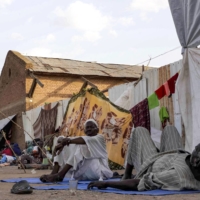 Men sit and lie outside shelters at the Hasahisa secondary school on Monday, which has been made into a make-shift camp to house people fleeing violence in war-torn Sudan. | AFP-JIJI
