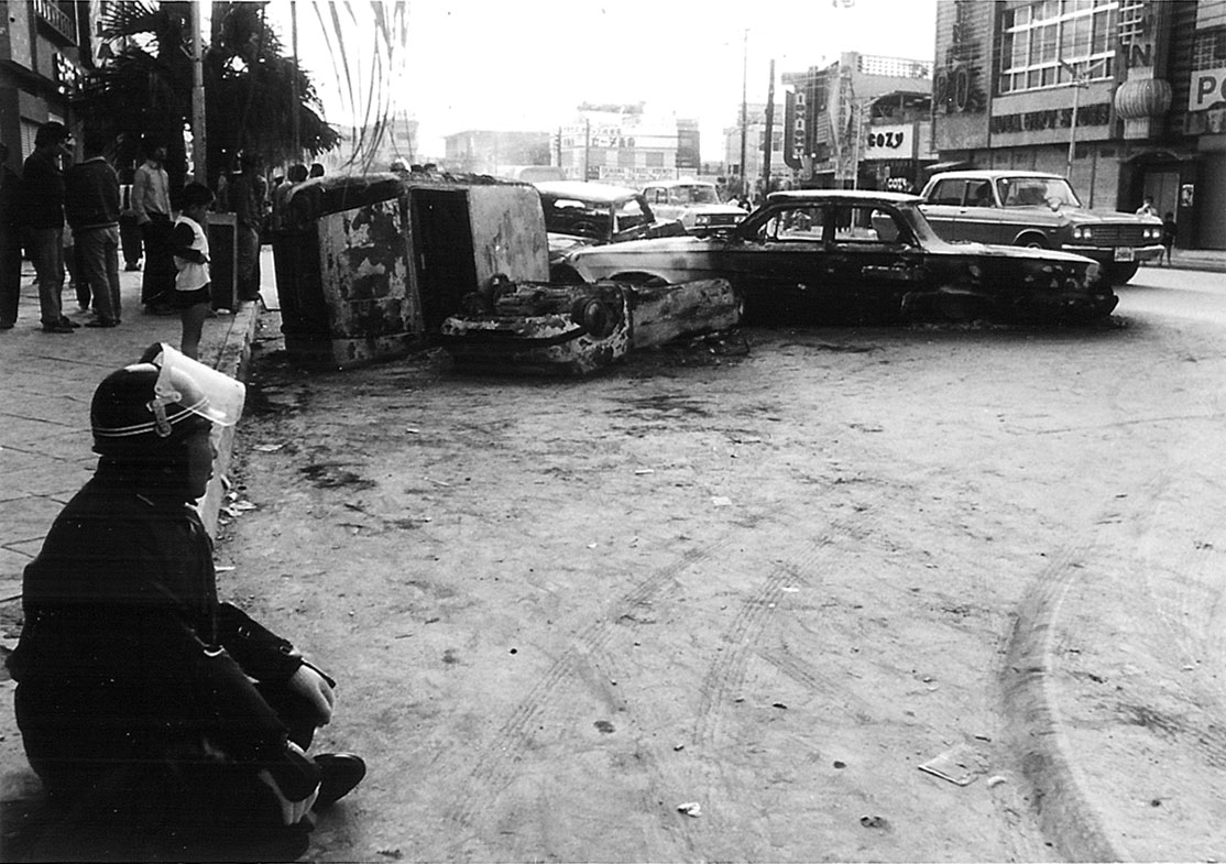 Aftermath of disturbances: As he sits on the curb, an Okinawan policeman surveys wreckage following rioting on Dec. 20, 1970, in what is now the city of Okinawa. | COURTESY OF LARRY GRAY