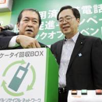 Insert here: Internal Affairs and Communications Minister Kunio Hatoyama (left) and Environment Minister Tetsuo Saito show off a mobile phone collection box in Tokyo Wednesday. | KYODO PHOTO