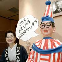 New shtick: Michiko Kakinoki, owner of famed Osaka eatery mannequin Kuidaore Taro, holds up a sign in the local dialect reading \"Please remember me\" as she tells reporters Monday the doll will now be used for promotional purposes. | KYODO PHOTO