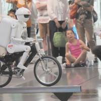 Murata Manufacturing Co.\'s Murata Seisaku-kun robot shows off his biking skills before an audience at the National Museum of Emerging Science and Innovation in Koto Ward, Tokyo, on Thursday. The robot rode the bike for 6 meters along a 2-cm-wide bar while maintaining its balance. | SATOKO KAWASAKI