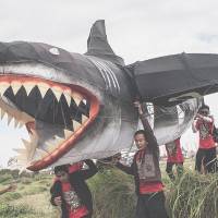 Participants taking part in the annual Bali Kite Festival carry a shark-shaped creation on the island on Friday. The seasonal gathering is intended to send a message to the gods to provide an abundant harvest. Approximately 1,120 traditional kites were launched into the sky during the three-day event. | GETTY/KYODO