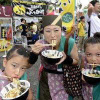 A woman and two girls Sunday eat \"udon\" noodles made in Okayama Prefecture, which won the recent U-1 Grand Prix noodle contest held in Yoyogi Park, Tokyo. Okayama\'s \"bukkake udon\" was the best-seller on the menu at the weekend event. | KYODO