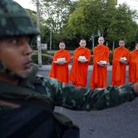 A Thai soldier stands guard as Buddhist monks beg for alms at a temple near Government House in Bangkok on Friday, a day after the army seized power in a coup. The military tightened its grip Sunday as it moved to douse smouldering protests fueled by social media and to rally commercial agencies and business to revitalize a battered economy. | REUTERS