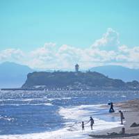People gather on a beach along the Kamakura coast, with Enoshima island in the background.  | \'ENOSHIMA ISLAND\' BY YOSHIKAZU TAKADA USED UNDER CC BY 2.0
