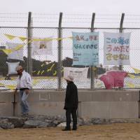 A couple stroll along the fence of U.S. Marine Corps Camp Schwab on the Henoko beach in Nago, Okinawa Prefecture. The fence bears anti-U.S. base protest banners. The banners read: \"Although U.S. Military is a menace, stop new base\" and \"We won\'t let you fill in this sea.\" | AP