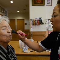 A care worker applies makeup to Ayako Sakai, a former nurse and housewife who was diagnosed with Alzheimer\'s disease almost 10 years ago, at the Soyokaze day care center in Osaka in August. | BLOOMBERG