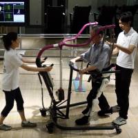 Tasuku Takahashi (center) uses the Lower Limb Model HAL (Hybrid Assistive Limb) device, manufactured by Cyberdyne. The 82-year-old stroke victim is paralyzed down his left side. He is seen during a HAL training session at the Cyberdyne studio in Tsukuba, Ibaraki Prefecture, on July 22. | REUTERS