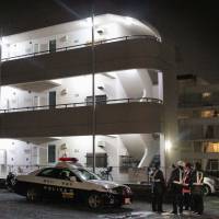 Investigators assemble Sunday night in front of an apartment in Atsugi, Kanagawa Prefecture, where an unidentified body was found wrapped in taped-together plastic bags. The residents of the unit were reported missing. | AP/MODIFIED BY THE JAPAN TIMES