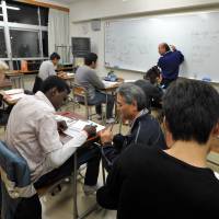 Hitoshi Ikeda (standing at the whiteboard) and Shuji Wachi (kneeling beside a desk) teach math to a class of nontraditional students at Bunka Junior High School on Dec. 10. | YOSHIAKI MIURA