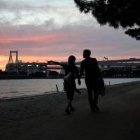 A couple walk in front of the Rainbow Bridge at dusk last August in Tokyo\'s Odaiba waterfront district. | BLOOMBERG