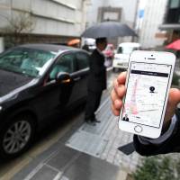 An Uber Japan Co. employee displays the smartphone app he has just used to call the driver (background) and car to the company\'s Tokyo office in March last year. Uber has suspended its pilot project in the city of Fukuoka after transport authorities objected, citing regulation breaches. | BLOOMBERG