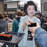 A geisha passes out boxes of sake in Kanazawa, Ishikawa Prefecture, on Saturday, as tourists descended on the scenic city, known for its traditional homes and Kenrokuen Garden, one of the country\'s \"three great gardens,\" after a new shinkansen line linking Kanazawa with Tokyo opened earlier in the day. | KYODO