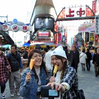 Nurses Chihiro Tanikai (left) and Mizuki Takaya take a selfie as they eat slices of pineapple on skewers in front of the entrance to the Ameyoko shopping arcade in March. | YOSHIAKI MIURA