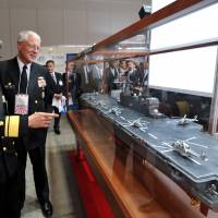 Maritime Self-Defense Force Rear Adm. Tadatsune Nishiyama (left) shows a model of the Izumo helicopter destroyer to U.S. Navy Capt. James Dick during the MAST Asia 2015 defense trade show at the Pacifico Yokoyama convention center on Wednesday. | YOSHIAKI MIURA