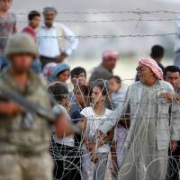 Syrian Kurds from Kobani wait behind the border fences to cross into Turkey as they are pictured from the Turkish border town of Suruc in Sanliurfa province, Turkey, Thursday after Islamic State fighters attacked the town, some allegedly wearing Kurdish uniforms and coming in from Turkey. | REUTERS