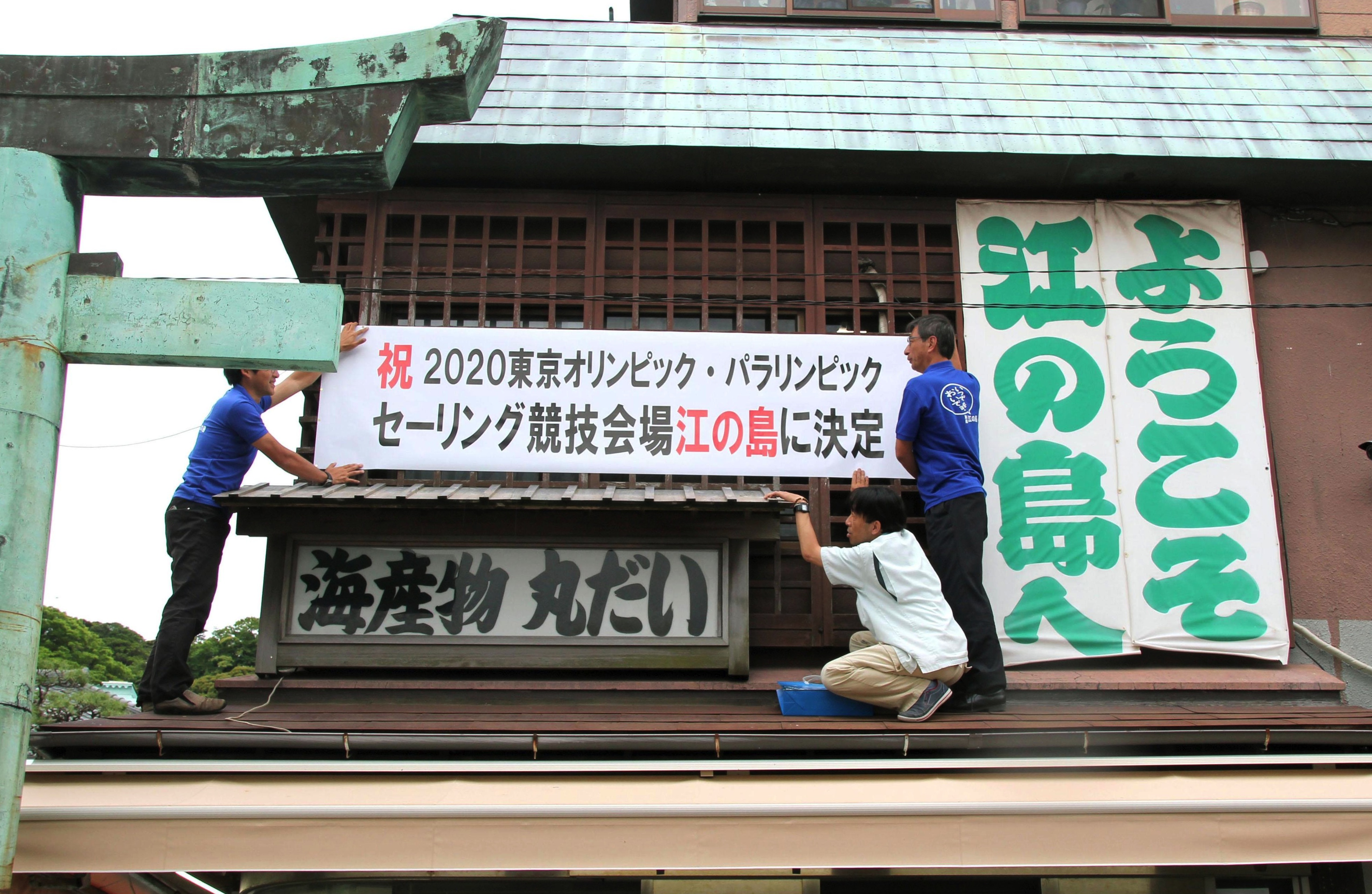 Workers hang a banner Tuesday on a building on Enoshima off Fujisawa, Kanagawa Prefecture, celebrating the island's choice to host the sailing events in the 2020 Tokyo Olympics. | KYODO