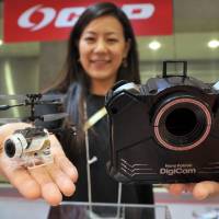 A CCP Co. employee shows off the Nano Falcon DigiCam, a remote-controlled copter that can shoot photos or videos from the air, at International Tokyo Toy Show 2015 at Tokyo Big Sight in Koto Ward on Thursday. The 20-gram drone can also be folded up and snapped into the cavity in the control unit to form a hand-held digital camera. | YOSHIAKI MIURA