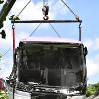 A tour bus that struck a dump truck and went off an expressway in Yokkaichi, Mie Prefecture, is lifted by crane from a tea plantation on Tuesday. | KYODO